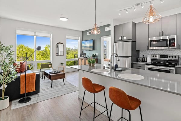 Modern open-plan kitchen and living room with stainless steel appliances, gray island, orange bar stools, and views of green landscape through large windows.